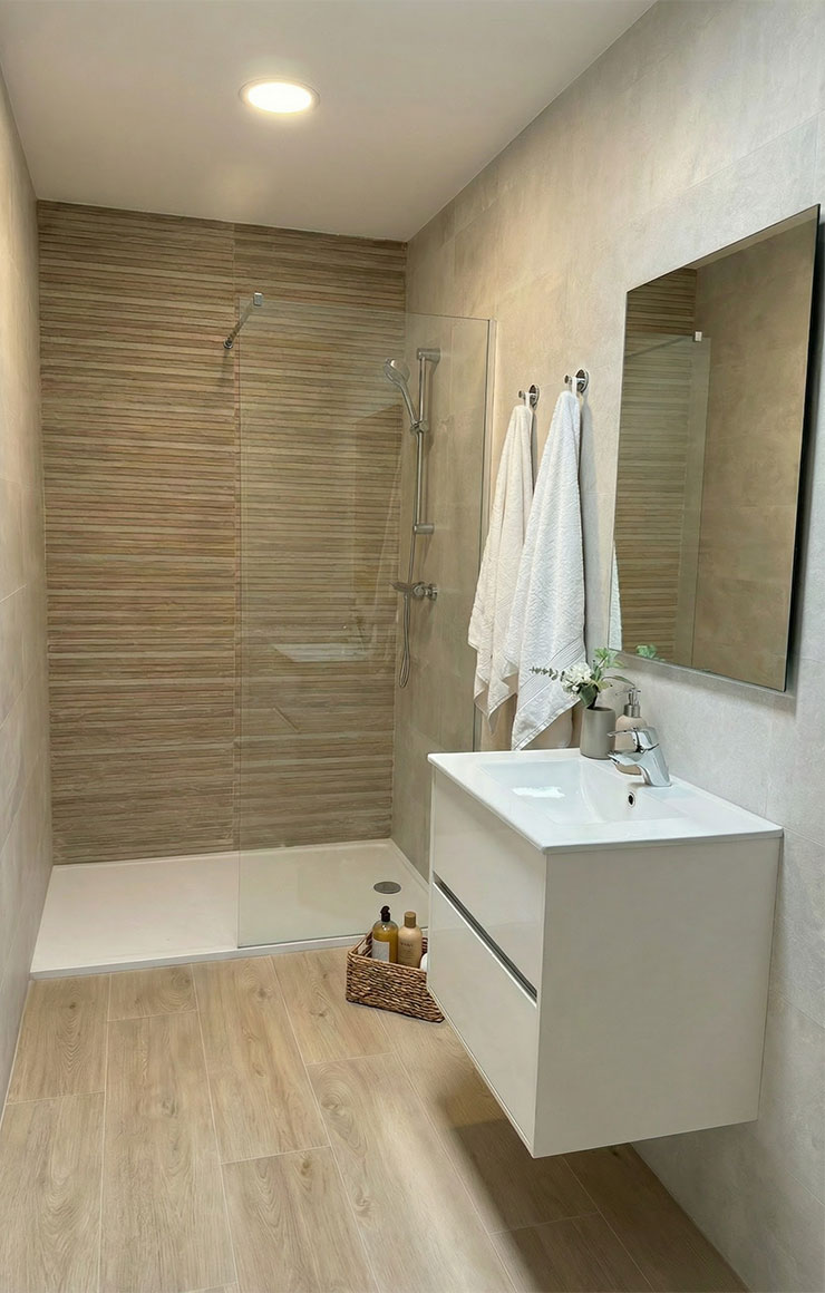 Cuarto de baño moderno con plato de ducha y mampara de perfilería negra. Cuenta con mueble de lavabo suspendido blanco, espejo amplio y acabados cerámicos imitación madera en chalet de Valdemoro.