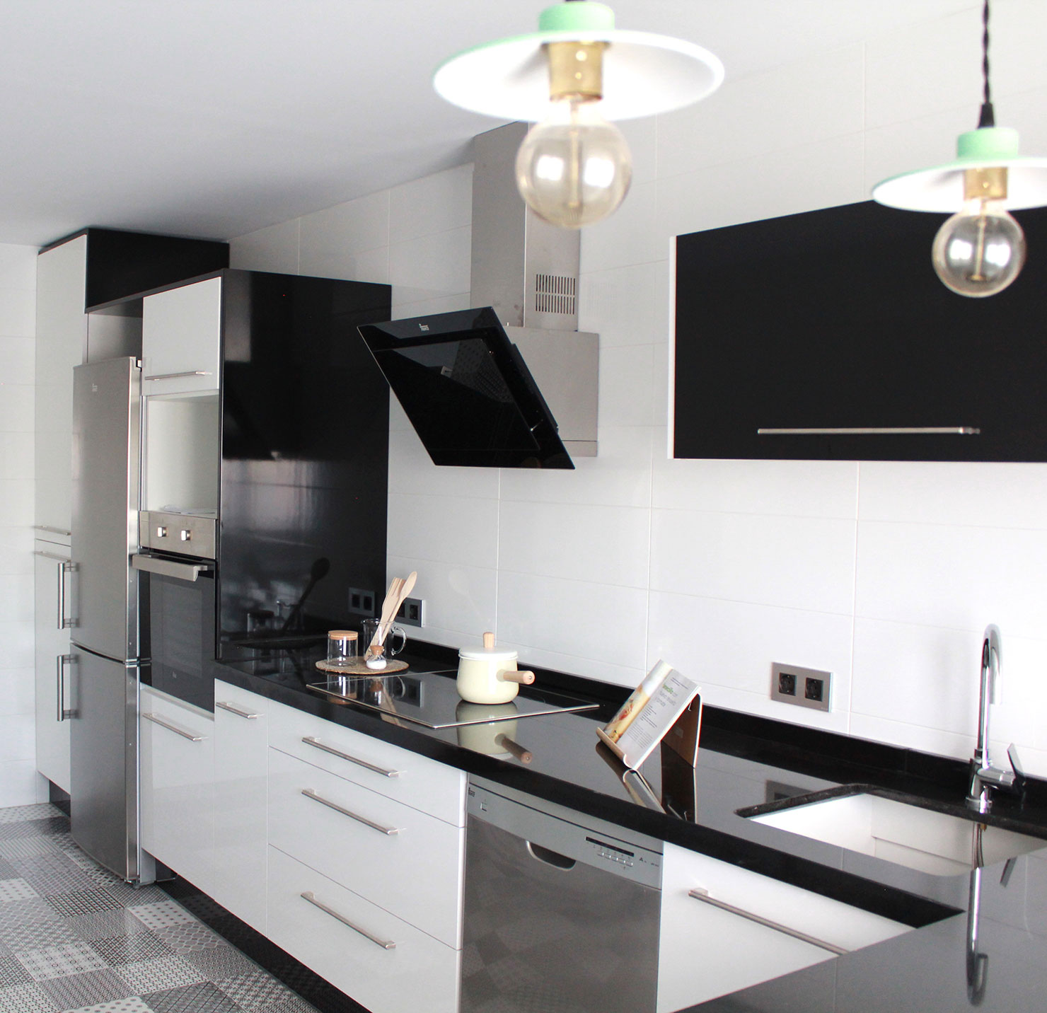 Cocina de estilo moderno en blanco y negro con encimera oscura y suelo de baldosas hidráulicas geométricas. Equipada con electrodomésticos de acero inoxidable, campana decorativa de cristal negro y lámparas industriales en chalet de Valdemoro.