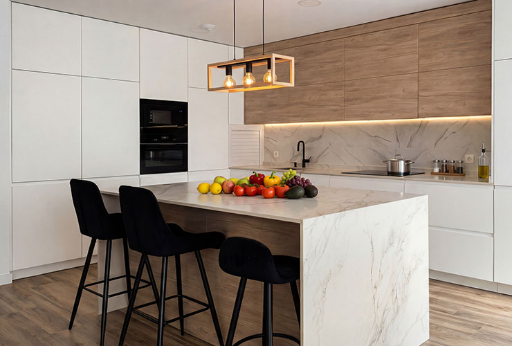 Diseño de cocina moderna en chalet de obra nueva en Valdemoro. Cuenta con una amplia isla central con barra de desayuno, combinación de armarios blancos y acabado madera, e iluminación LED integrada bajo mueble.